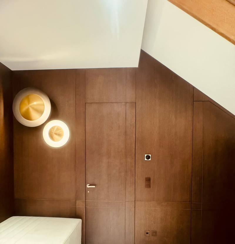 Mur habillé de panneaux en bois foncé sous pente, porte affleurante intégrée, deux appliques murales rondes dorées et banquette beige
