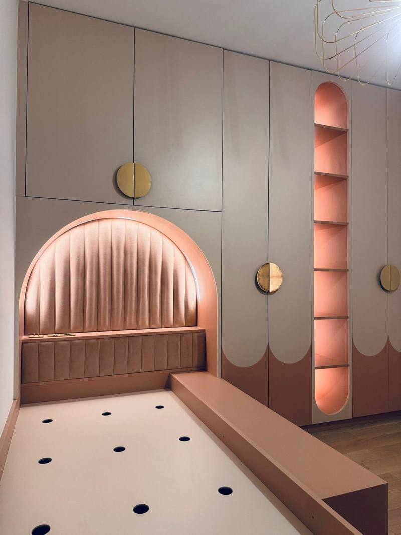 Chambre d'enfant avec armoires toute hauteur grège et rose, tête de lit capitonnée en arche éclairée par LED, niche à étagères lumineuse et poignées dorées