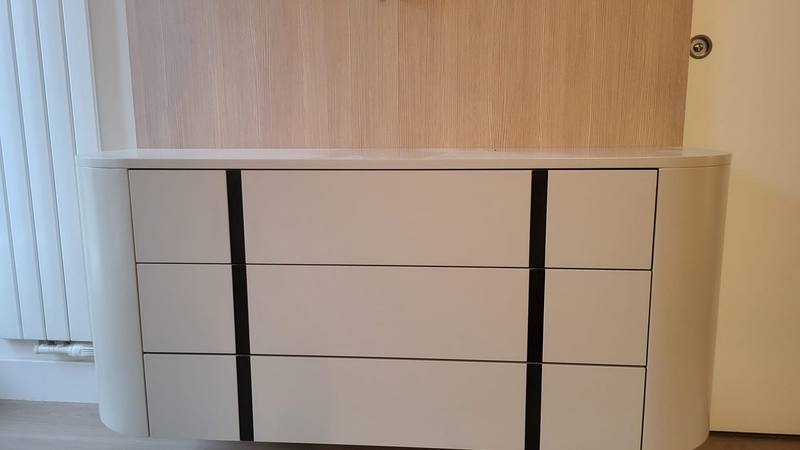 Commode suspendue laquée beige clair à angles arrondis, neuf façades de tiroirs rythmées par deux bandes noires verticales, adossée à un panneau mural en bois clair