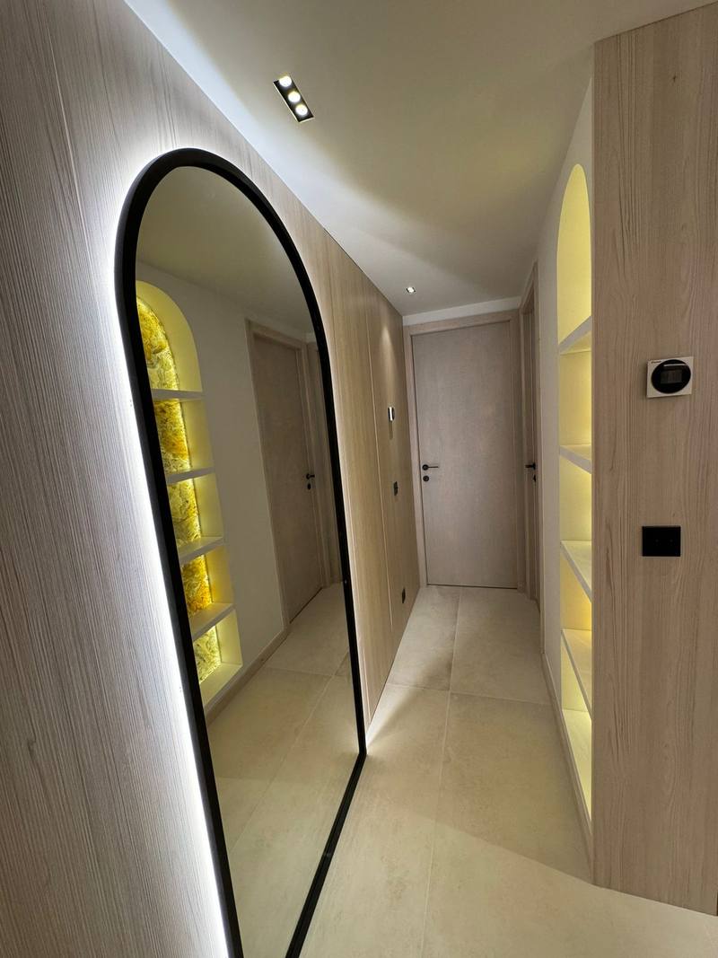 Couloir étroit aux murs en bois clair, grand miroir en arche à liseré lumineux, niche à étagères éclairées en face, portes claires et sol carrelé