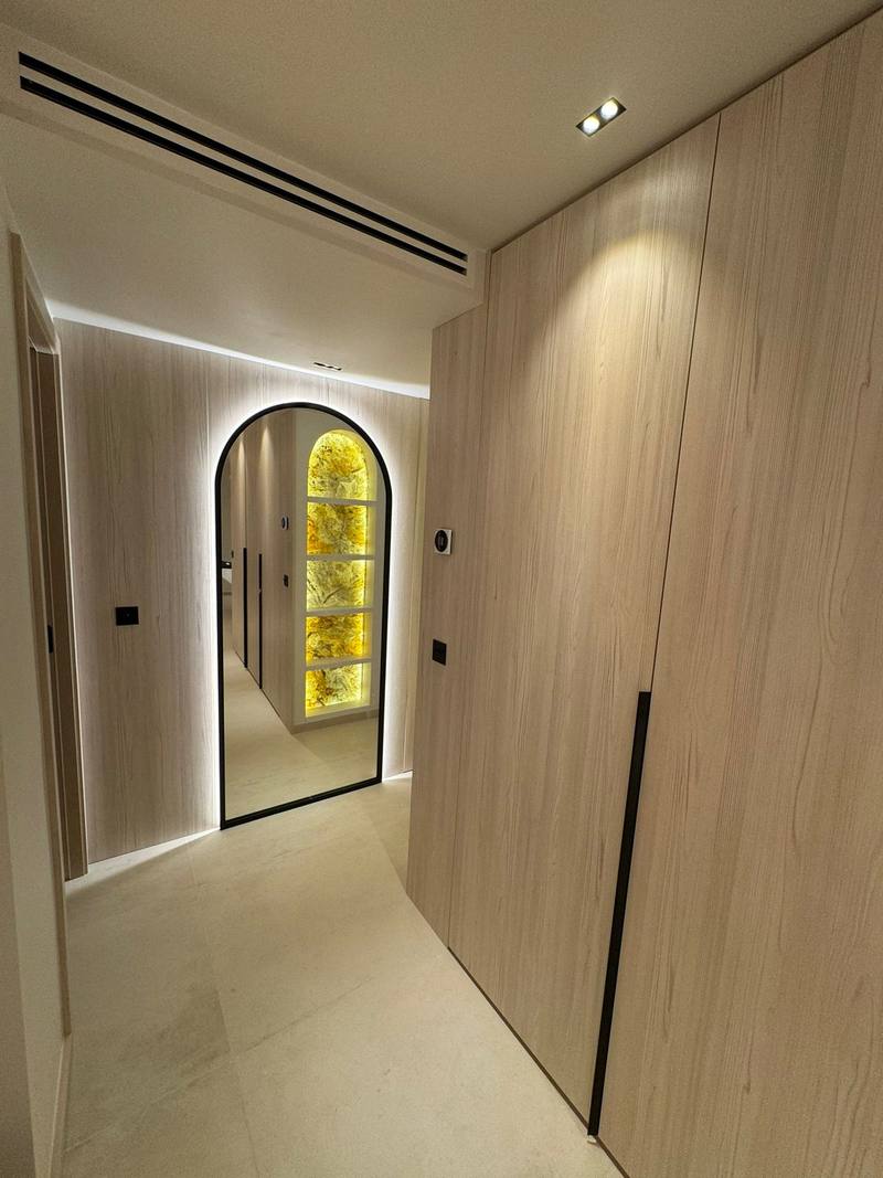 Couloir habillé de panneaux bois clair à poignées noires intégrées, miroir en arche cerclé de noir et rétroéclairé, sol en grands carreaux clairs