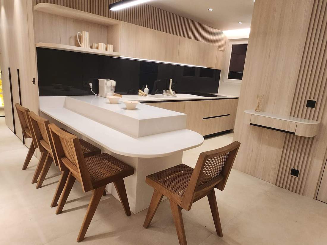 Cuisine en bois clair avec tasseaux, crédence en verre noir, bar blanc à bout arrondi entouré de chaises en bois et cannage, suspension linéaire noire