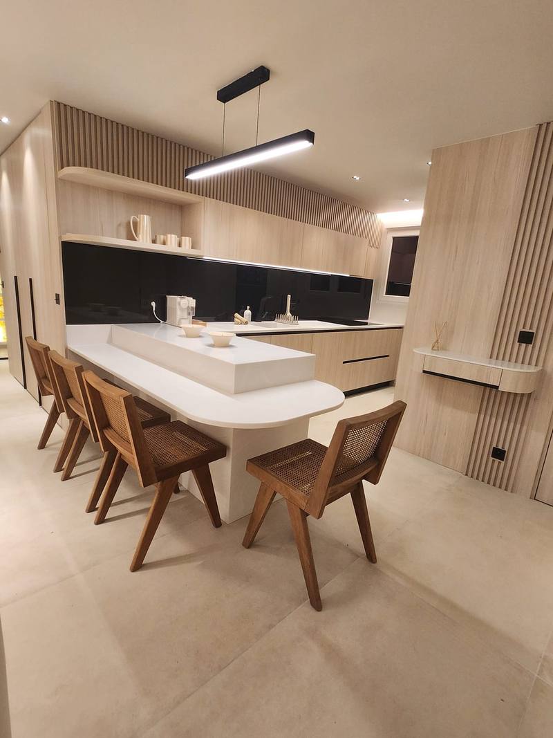 Cuisine en bois clair avec tasseaux, crédence en verre noir, bar blanc à bout arrondi entouré de chaises en bois et cannage, suspension linéaire noire