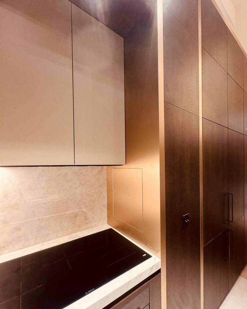 Cuisine avec colonnes en bois foncé à filets bronze, placards hauts laqués beige, crédence et plan en pierre claire veinée, plaque induction