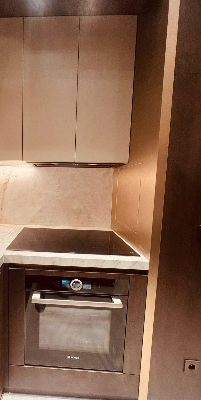 Cuisine en L avec façades hautes beige mat sans poignée, plan et crédence en pierre claire veinée d'ocre, plaque à induction et four encastré