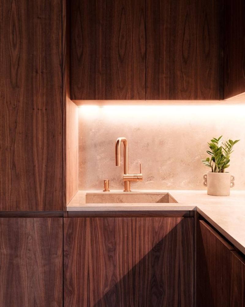 Angle de cuisine en bois foncé veiné, évier encastré et plan en pierre claire, crédence pierre éclairée par LED, robinet cuivre