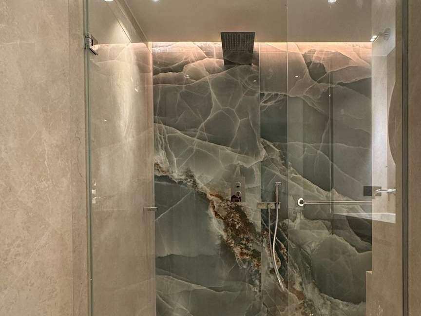 Douche à l'italienne vitrée dans une salle de bain en grands carreaux beige veinés, mur du fond effet onyx vert, caniveau de sol, ciel de pluie encastré et gorge lumineuse