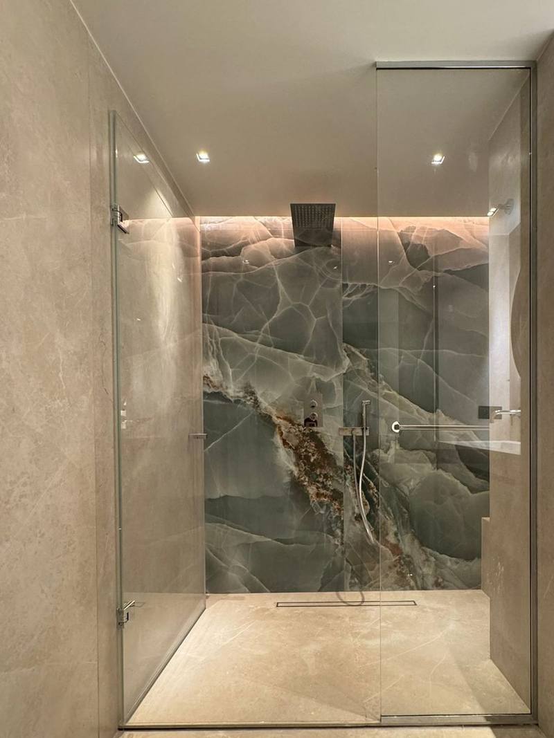 Douche à l'italienne vitrée dans une salle de bain en grands carreaux beige veinés, mur du fond effet onyx vert, caniveau de sol, ciel de pluie encastré et gorge lumineuse