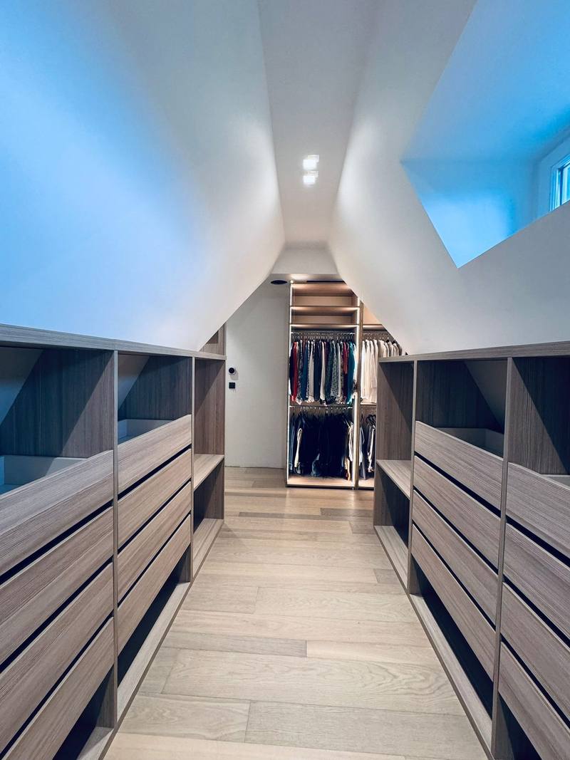 Dressing sous combles avec deux rangées de commodes à tiroirs et niches en bois, allée en parquet clair et penderies éclairées au fond