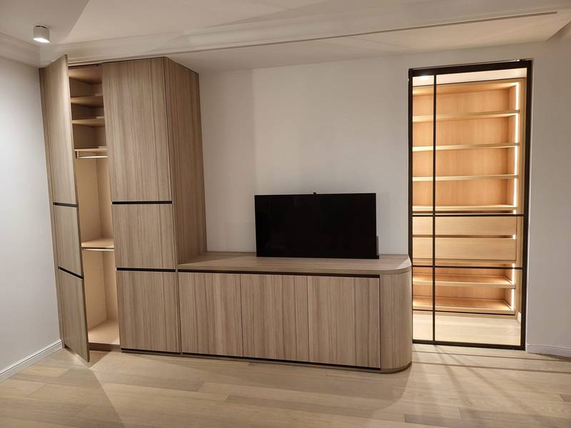Mur de rangements en bois clair avec meuble TV bas à angle arrondi, armoire ouverte sur penderie et verrière à profilés noirs devant un dressing éclairé