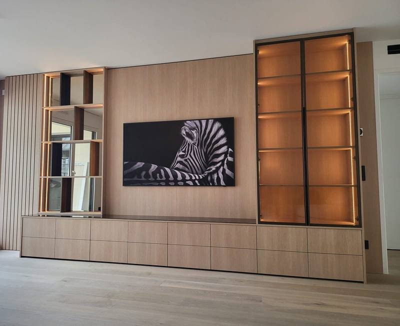 Mur de salon en bois clair avec meuble TV bas à tiroirs, étagères miroir et vitrine à portes vitrées encadrées de métal, éclairées par LED