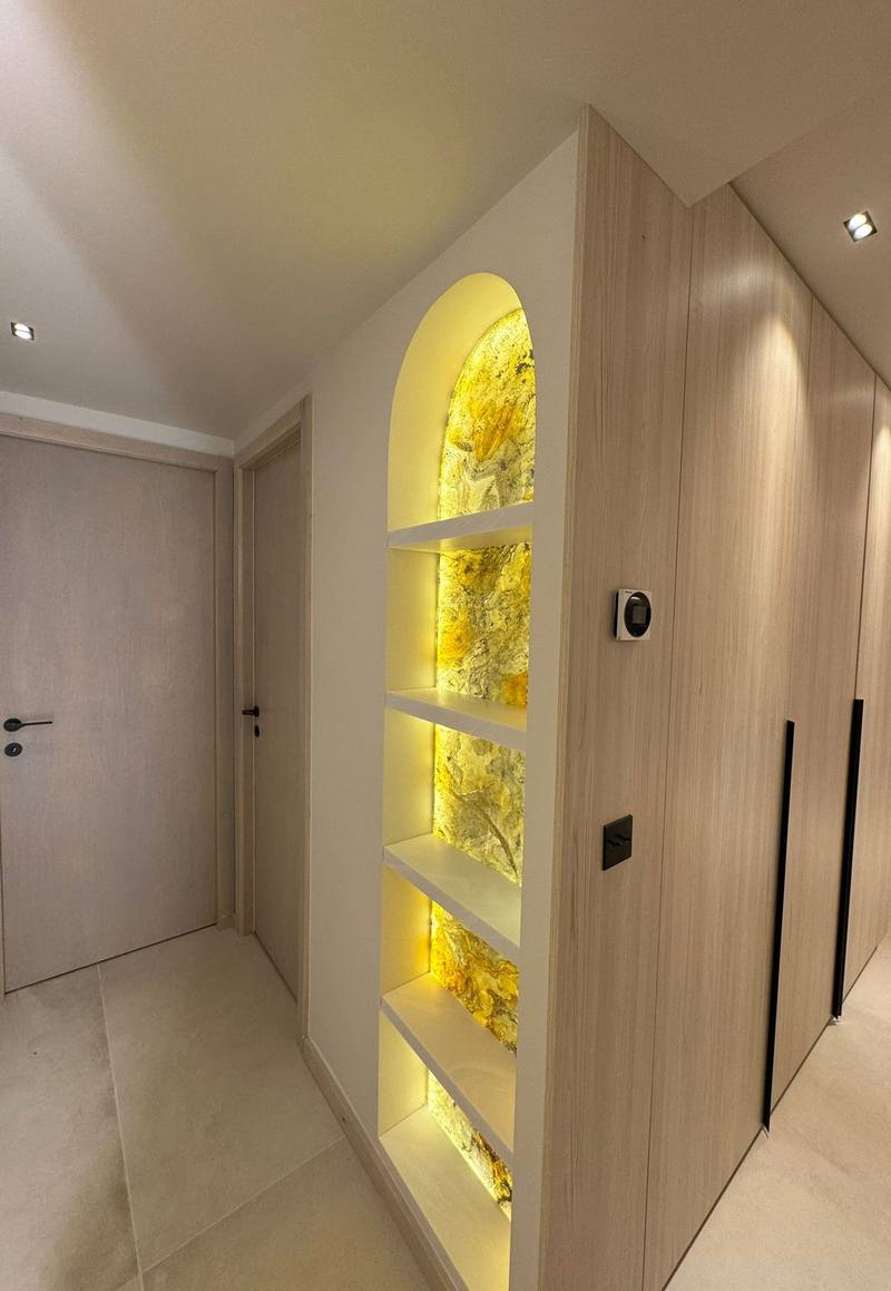 Niche en arche blanche à étagères, fond en pierre translucide jaune rétroéclairée, à côté de placards toute hauteur en bois clair à poignées noires