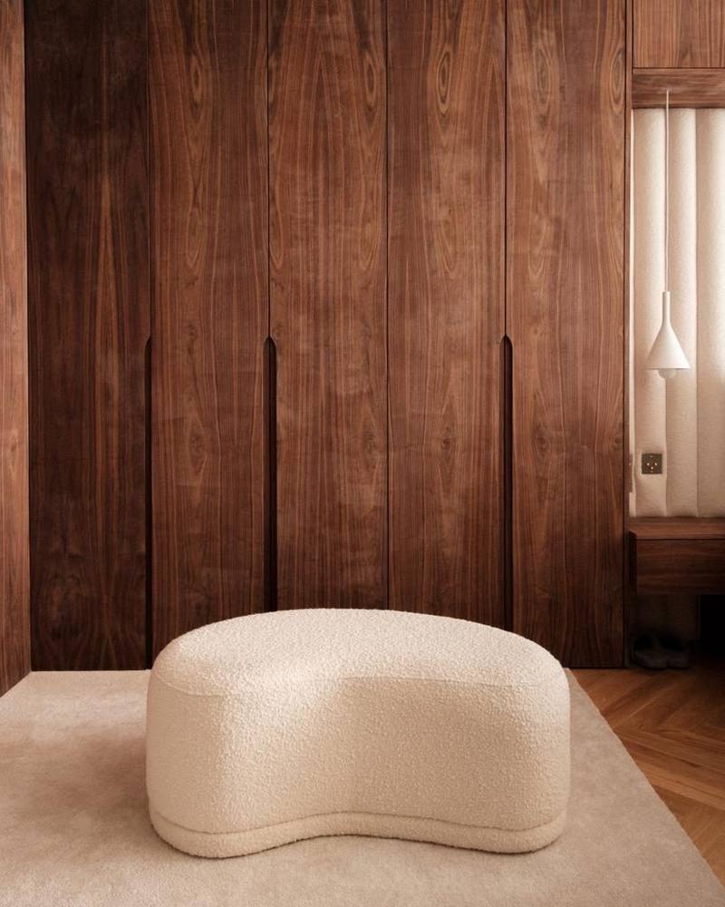 Mur de placards toute hauteur en bois foncé veiné à poignées encastrées, pouf bouclette crème sur tapis, chevet et tête de lit capitonnée à droite