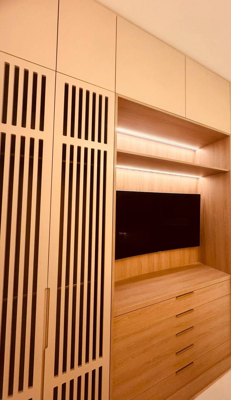 Mur de placards laqués beige à portes ajourées verticales, niche centrale en bois clair avec télévision, étagères éclairées par LED et commode