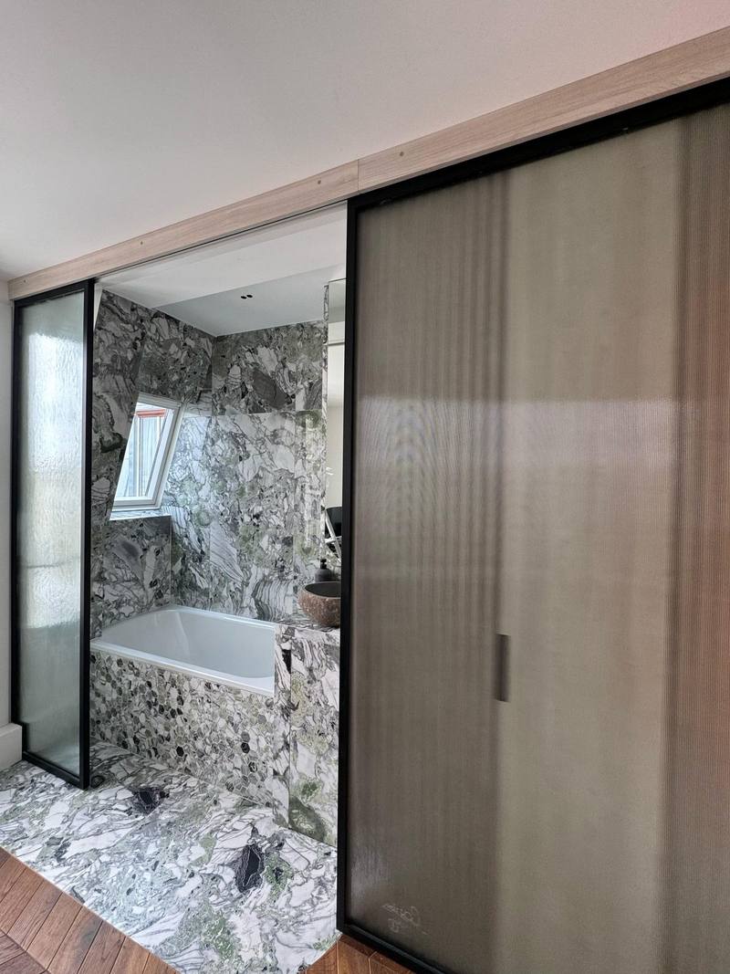 Salle de bain en marbre vert et blanc avec baignoire, tablier en mosaïque hexagonale, vasque en pierre, cloison et porte coulissante en verre cannelé