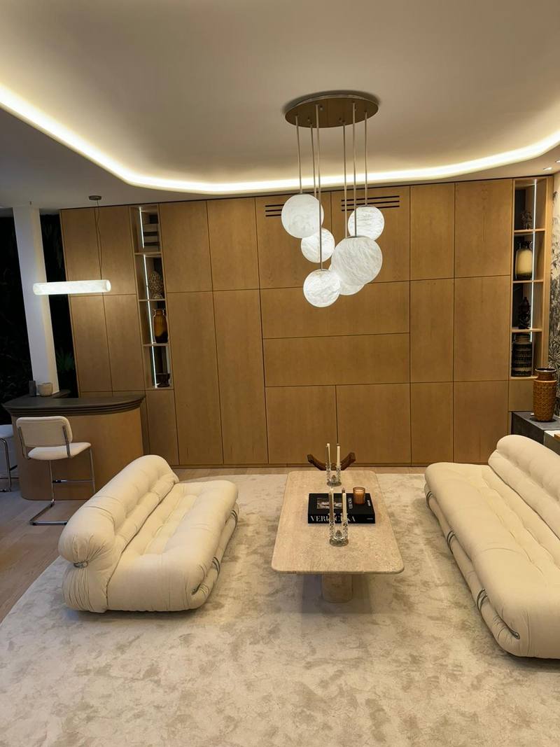 Salon avec mur de placards en bois naturel encadrant des niches éclairées, plafond à gorge lumineuse, suspension à globes blancs, canapés crème
