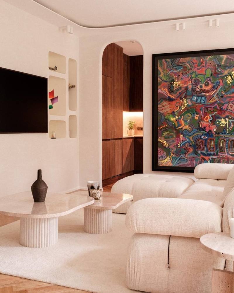 Salon clair aux lignes arrondies : niches murales près de la télévision, passage en arche vers une cuisine en bois foncé, tables basses en pierre, canapé crème et grand tableau coloré