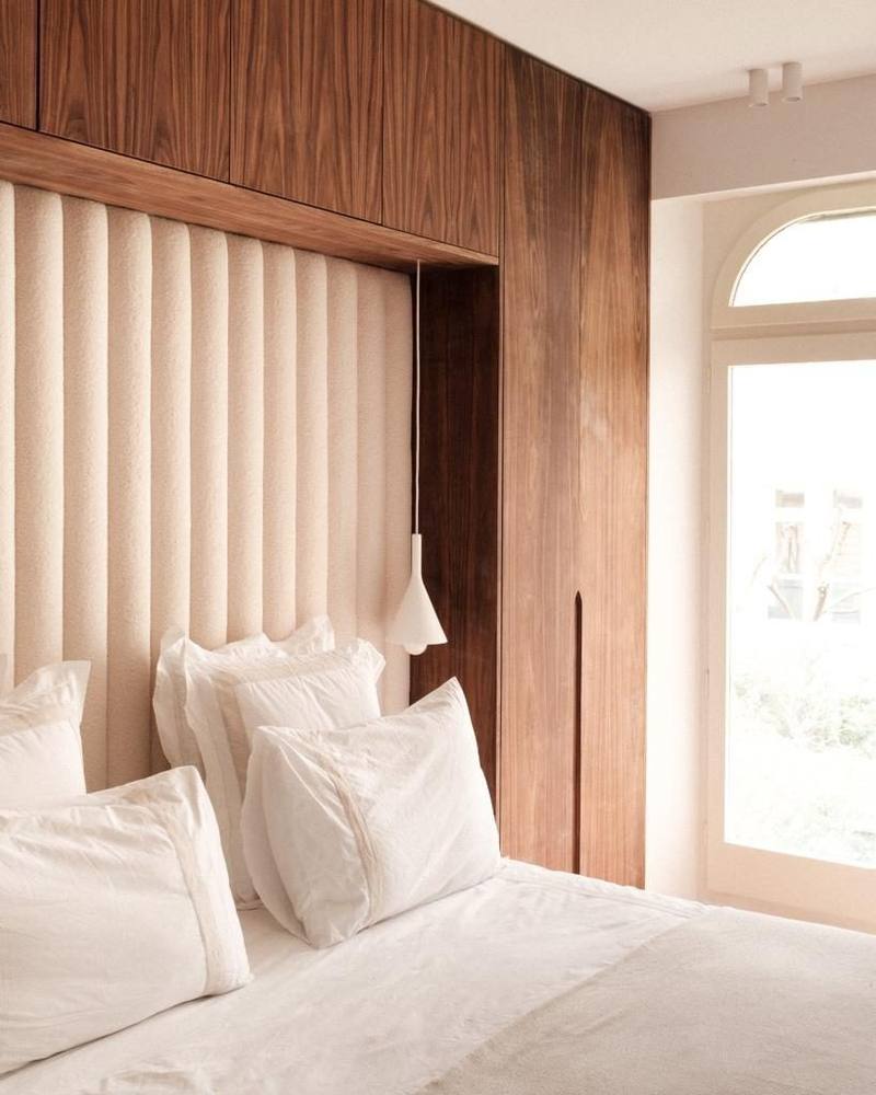 Chambre avec tête de lit capitonnée en bouclette crème encadrée de placards en bois foncé veiné, suspension blanche et fenêtre cintrée lumineuse