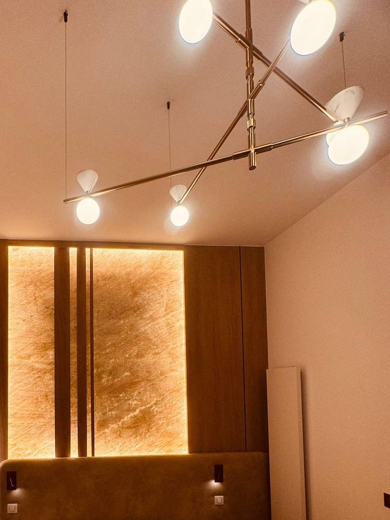Chambre avec mur de tête de lit en bois, panneau de pierre translucide rétroéclairé, tête de lit en tissu à liseuses intégrées, radiateur vertical blanc et suspension dorée à globes