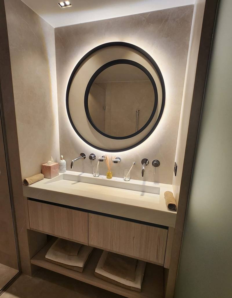 Plan vasque monobloc blanc, robinetterie murale chromée double, miroir rond noir rétroéclairé LED, meuble bois clair à deux tiroirs et niche ouverte