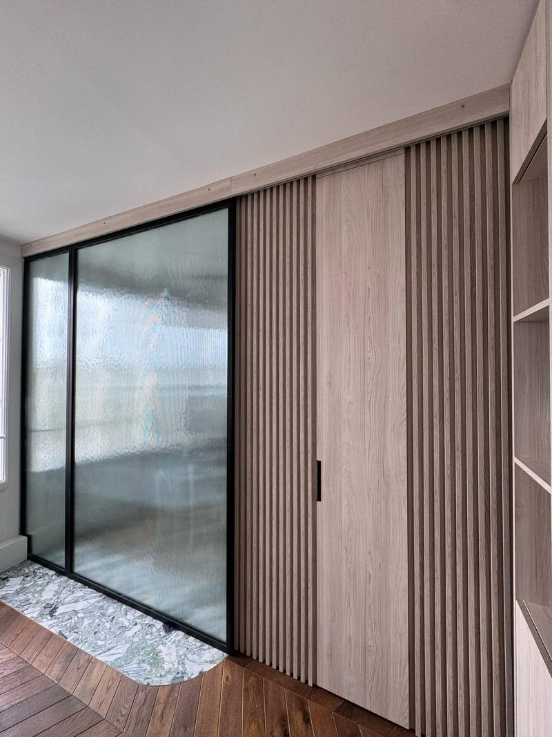 Cloison vitrée cannelée à cadre noir, mur de tasseaux en bois clair intégrant une porte affleurante, seuil en marbre vert, parquet en chevrons